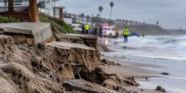 Close-up of storm-damaged coastal sand and erosion near a Southern California beach, with blurred emergency activity in the background.