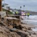 Close-up of storm-damaged coastal sand and erosion near a Southern California beach, with blurred emergency activity in the background.
