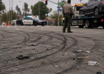 Close-up of tire marks and scattered debris across an intersection, with a blurred deputy and impounded vehicle visible in the background.