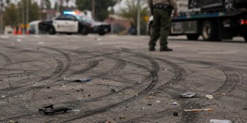 Close-up of tire marks and scattered debris across an intersection, with a blurred deputy and impounded vehicle visible in the background.