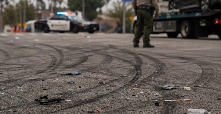 Close-up of tire marks and scattered debris across an intersection, with a blurred deputy and impounded vehicle visible in the background.