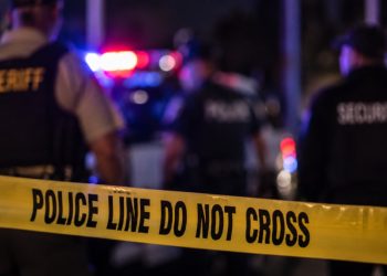 Close-up of yellow police barrier tape with blurred emergency lights, officers and security personnel in the background, illustrating a Malibu trespassing investigation.
