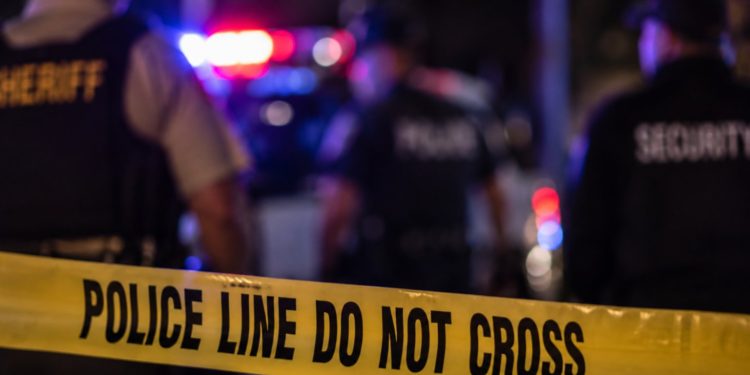 Close-up of yellow police barrier tape with blurred emergency lights, officers and security personnel in the background, illustrating a Malibu trespassing investigation.