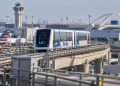 LAX SkyLink train on its elevated guideway near the airport terminals, with testing infrastructure and airport facilities visible in the background.