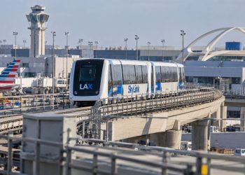 LAX SkyLink train on its elevated guideway near the airport terminals, with testing infrastructure and airport facilities visible in the background.
