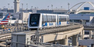LAX SkyLink train on its elevated guideway near the airport terminals, with testing infrastructure and airport facilities visible in the background.