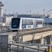 LAX SkyLink train on its elevated guideway near the airport terminals, with testing infrastructure and airport facilities visible in the background.