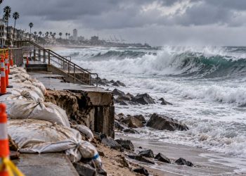 Large waves approaching the Long Beach Peninsula under overcast skies, with a damaged coastal barrier and emergency preparations visible near the shoreline.