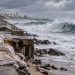 Large waves approaching the Long Beach Peninsula under overcast skies, with a damaged coastal barrier and emergency preparations visible near the shoreline.