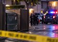 Police investigation scene near a Riverside apartment complex, with a dumpster enclosure and emergency vehicles blurred in the background.