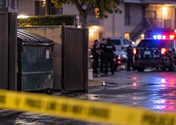Police investigation scene near a Riverside apartment complex, with a dumpster enclosure and emergency vehicles blurred in the background.
