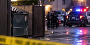 Police investigation scene near a Riverside apartment complex, with a dumpster enclosure and emergency vehicles blurred in the background.