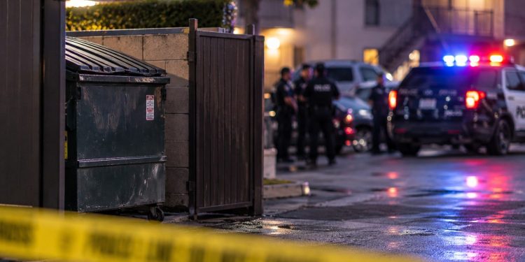 Police investigation scene near a Riverside apartment complex, with a dumpster enclosure and emergency vehicles blurred in the background.