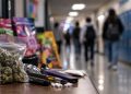 Close-up of assorted colorful drug packages and vaping devices on a table, with a heavily blurred school hallway and students in the background, illustrating an alleged narcotics operation targeting young students in Ventura County.