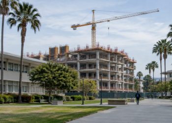 A concrete-framed building under construction beside older classroom buildings on a Los Angeles community college campus in afternoon daylight.