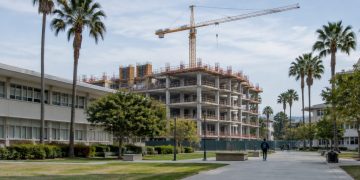 A concrete-framed building under construction beside older classroom buildings on a Los Angeles community college campus in afternoon daylight.