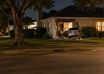 A one-story stucco house on a residential street in Sherman Oaks with a damaged exterior wall, seen from across the street at night under street lamps.