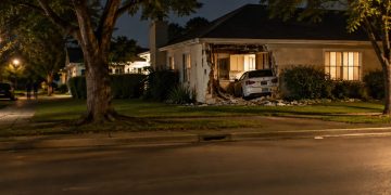 A one-story stucco house on a residential street in Sherman Oaks with a damaged exterior wall, seen from across the street at night under street lamps.