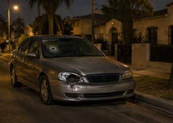 A parked sedan with a cracked windshield and broken headlight on a residential street of stucco homes and palm trees in Westmont at night.