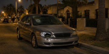 A parked sedan with a cracked windshield and broken headlight on a residential street of stucco homes and palm trees in Westmont at night.