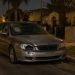 A parked sedan with a cracked windshield and broken headlight on a residential street of stucco homes and palm trees in Westmont at night.