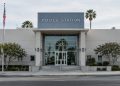 A police station building in Van Nuys in neutral daylight, its institutional facade sharp and centered, palm trees and neighborhood buildings blurred behind.