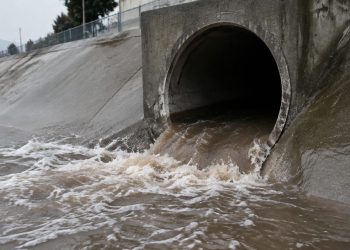 A sewer tunnel outlet in a San Bernardino flood-control channel wall, with brown surging water at its opening on a morning under overcast sky.