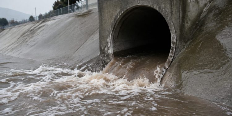 A sewer tunnel outlet in a San Bernardino flood-control channel wall, with brown surging water at its opening on a morning under overcast sky.