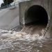 A sewer tunnel outlet in a San Bernardino flood-control channel wall, with brown surging water at its opening on a morning under overcast sky.