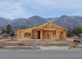 A wood-framed house under construction on a cleared residential lot in Altadena, with empty lots and the San Gabriel Mountains behind in daylight.