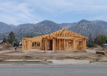 A wood-framed house under construction on a cleared residential lot in Altadena, with empty lots and the San Gabriel Mountains behind in daylight.