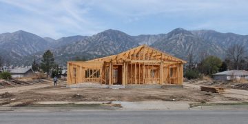 A wood-framed house under construction on a cleared residential lot in Altadena, with empty lots and the San Gabriel Mountains behind in daylight.