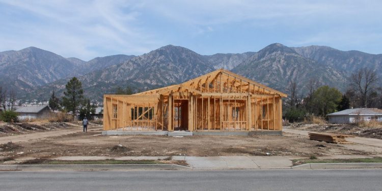 A wood-framed house under construction on a cleared residential lot in Altadena, with empty lots and the San Gabriel Mountains behind in daylight.