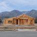 A wood-framed house under construction on a cleared residential lot in Altadena, with empty lots and the San Gabriel Mountains behind in daylight.