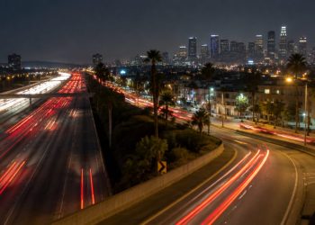 Aerial view of a multilane freeway at night in South Los Angeles with an off-ramp curving toward a boulevard lined with palm trees and low-rise buildings.
