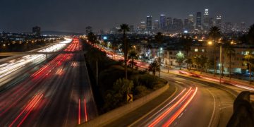 Aerial view of a multilane freeway at night in South Los Angeles with an off-ramp curving toward a boulevard lined with palm trees and low-rise buildings.