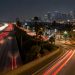 Aerial view of a multilane freeway at night in South Los Angeles with an off-ramp curving toward a boulevard lined with palm trees and low-rise buildings.