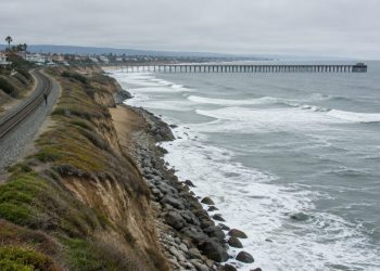 Aerial view of a single railroad track along a beachfront bluff in San Clemente, with waves breaking near the rocky slope below the rails in daylight.