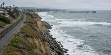Aerial view of a single railroad track along a beachfront bluff in San Clemente, with waves breaking near the rocky slope below the rails in daylight.
