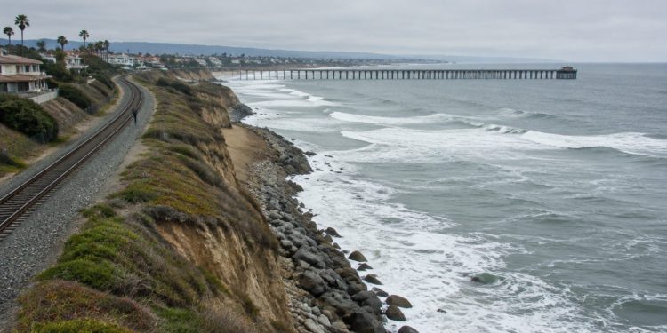 Aerial view of a single railroad track along a beachfront bluff in San Clemente, with waves breaking near the rocky slope below the rails in daylight.