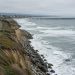Aerial view of a single railroad track along a beachfront bluff in San Clemente, with waves breaking near the rocky slope below the rails in daylight.