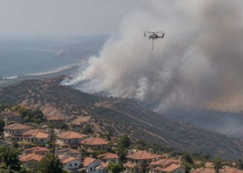 Aerial view of a smoke plume rising over dry hills above suburban homes in San Clemente, with a firefighting helicopter in flight in afternoon daylight.