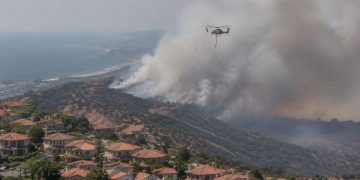 Aerial view of a smoke plume rising over dry hills above suburban homes in San Clemente, with a firefighting helicopter in flight in afternoon daylight.