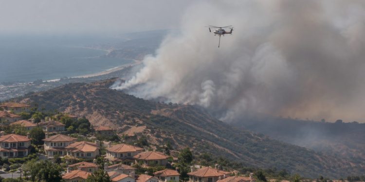 Aerial view of a smoke plume rising over dry hills above suburban homes in San Clemente, with a firefighting helicopter in flight in afternoon daylight.