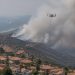 Aerial view of a smoke plume rising over dry hills above suburban homes in San Clemente, with a firefighting helicopter in flight in afternoon daylight.