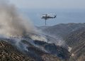 Aerial view of smoke rising over dry chaparral hills in the Santa Monica Mountains near Malibu as a firefighting helicopter flies in afternoon daylight.