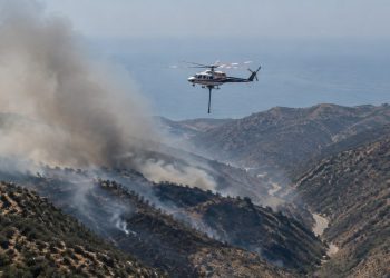 Aerial view of smoke rising over dry chaparral hills in the Santa Monica Mountains near Malibu as a firefighting helicopter flies in afternoon daylight.