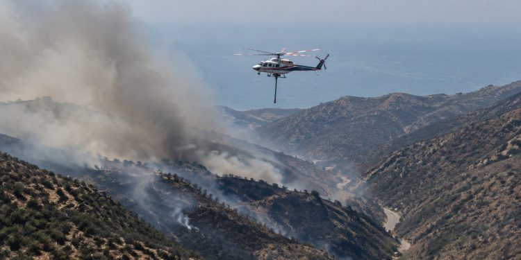 Aerial view of smoke rising over dry chaparral hills in the Santa Monica Mountains near Malibu as a firefighting helicopter flies in afternoon daylight.