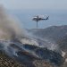 Aerial view of smoke rising over dry chaparral hills in the Santa Monica Mountains near Malibu as a firefighting helicopter flies in afternoon daylight.