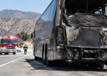 Charred rear of a tour bus on the shoulder of a freeway in Burbank in morning daylight, with a fire engine blurred in the background.